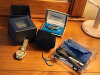 Photo displaying all items on wooden surface including 3 wristwatches, one in a Hamilton box, and a plastic case with watch tools.