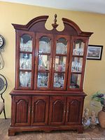 Front view of the large wooden curio cabinet showing the upper glass doors with visible glassware and china inside, lower closed wooden doors, and ornate top.
