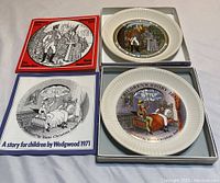 Two Wedgwood children's story plates placed next to their original colorful story boxes featuring on-cover illustrations. Plates show detailed color scenes from Hans Christian Andersen stories with fluted rims.