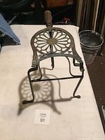 Photo showing angled view of antique cast iron trivet or stand with decorative top and wooden handle, highlighting structure and design