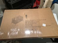 Boxed baby crib wrapped in plastic film with an illustration of assembled crib on box top.