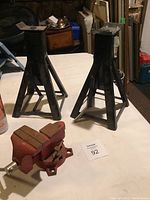 Photo showing two black metal jack stands and one red bench vise on a white table surface.