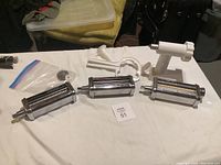 Lot of kitchen accessories including three stainless steel pasta rollers and a white plastic clamp on a white tablecloth.