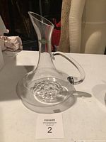 Photo of the clear glass decanter with handle on a white table, showing its shape and bottom design.
