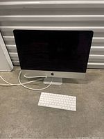 Front view of Apple iMac computer showing screen and keyboard on floor with power cable attached.