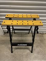 Front view of Husky work bench showing wooden top with multiple holes and measurement markings, black metal legs, and Husky branding.