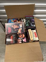 Open cardboard box filled with various VHS tapes in original cases, showing titles including 'Passenger 57,' 'Alien Nation,' and 'Scanners'.