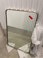 Front view of the rectangular wall mirror with silver metal frame leaning against a corrugated metal wall, partially wrapped in protective packing material.