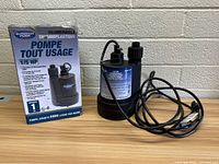 Image showing the submersible pump and packaging together on a surface against a brick wall, product visible includes pump with attached power cord and original box showing bilingual labeling.