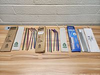 Three sets of stainless steel straws lying side by side, each with their box, fabric pouch, and cleaning brush.