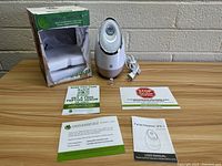 Wide angle view of Airome Facial Steamer SPA+ with original box, power cord, user manual, and promotional flyers on wooden table against pale cinderblock wall.