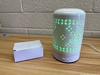 Photo showing diffuser with green light on, decorative cutout pattern visible, and small white box placed next to it on table