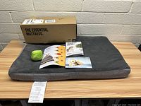 Photo of medium Bark! memory foam dog bed with associated box, plush green ball toy, and paperwork on a wooden table against a white brick wall.