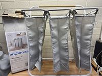 Front view of the rolling laundry hamper showing three beige fabric bags hanging from the white metal frame with wooden handle, box displayed