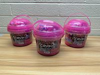 Three pink buckets of Compound Kings Butter Cloudz slime placed on a wooden surface against a brick wall background.
