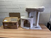 Left angled view of the beige Feandrea cat tree next to its box on wooden surface in front of light-colored brick wall.