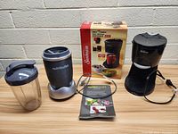 Overview of the Sunbeam Hot Shot Hot Water Dispenser with box, NutriBullet blender base and blending cup, plus user manual on a tiled floor.