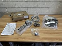 Full set of shower components including large round overhead rainfall shower head, handheld shower head, flexible hose, connector, and instructions displayed on wooden table.