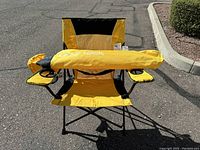 Yellow and black Kijaro folding chair unfolded outdoors with carrying bag resting across seat and armrests. Chair has dual cup holders on armrests and a breathable mesh back.