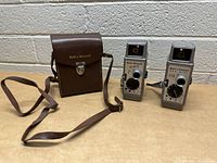 Two Bell & Howell Two Twenty 8mm cameras with square viewfinders next to a brown Bell & Howell carrying case with strap and clasp.