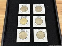 Six coins displayed in protective holders with handwritten labels identifying each coin by the site or landmark it features in Greece.