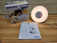 Philips Wake-Up Light operational with soft glow, digital time visible, original box, and manual shown on wooden table.
