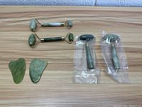 Top-down view of two jade facial rollers with gold accents, two gua sha stones, and two micro-needling facial massagers in plastic packaging on wooden surface.