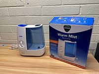 Vicks Warm Mist Humidifier with original box placed on wooden surface against brick wall.