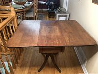 Front angle of antique drop leaf wooden table with two drop leaves open and central drawer visible.