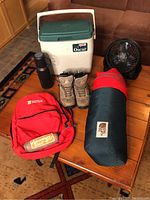 Full lot image showing hiking boots, sleeping bag, cooler, thermos, fan, and backpack on a table
