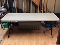 Full view of rectangular white plastic table with black folding legs in a basement setting.