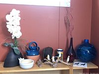 Photo showing a variety of home decor items including an artificial orchid arrangement, pottery jug and vases, decorative birds, and glass vases on wooden shelves against a red wall.