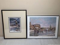 Photo showing two framed prints side by side on a white surface. Left is a smaller piece with a child and beach scene, right is a larger urban scene with people and buildings.