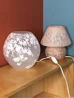 Two small glass lamps displayed on a wooden shelf against a blue and brown wall. The first is frosted glass with white floral patterns, the second is a rose pink and white speckled mushroom lamp with cord and switch.