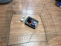 Metal wire pet pen arranged in square with small gate, placed on wood floor, includes a plastic container with pet grooming tools and a small white box.