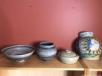 Four assorted pottery pieces including a large grey bowl, small marbled vase, beige lidded jar, and colorful sun and moon vase.