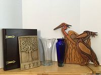 Photo showing all items in the lot including the wooden book box, the Tree of Life stone plaque, two wooden cranes sculptures, and two glass vases (clear and cobalt blue) placed side by side.