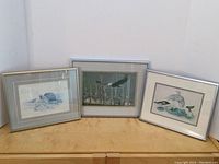 Three framed art prints displayed together on a wooden surface, depicting wildlife and indigenous village scenes.