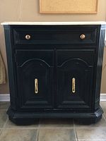 Front view of black Stanley cabinet showing single drawer with round gold knob, two doors with gold pulls, shaping details on door panels and front base.