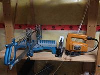Photo showing the blue Mastercraft miter box with attached saw, the Black & Decker orange jigsaw, and the black Mastercraft swivel saw on a wooden shelf.