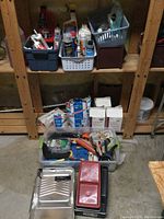 Shelf with plastic bins containing various tubes of caulking and silicone, paint brushes, rollers, small tools, and two paint trays with red and silver paint grids.