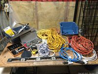 Wide shot showing the assortment of tools and cords on a wooden table in a workshop environment including ratchet set, level, extension cords, tape measure, and various hardware parts.