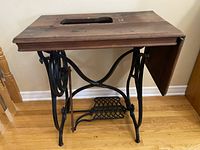 Front view of antique sewing machine table showing wooden top with cutout and foldable extension wing, cast iron ornate base with treadle pedal.