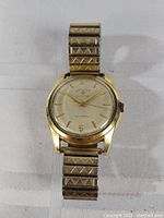 Full front view of gold-tone wristwatch with cream dial, gold hour markers and 'Grand Prix Election' branding, showing overall watch face and expandable bracelet.