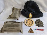 Photo shows two Canadian Forces knit caps, wooden emblem, metal insignias, ribbon bar, and two sealed MRE ration packs.