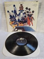 Photo showing the vinyl record partially out of the sleeve, disc is shiny and clean with label visible on black vinyl. Sleeve front shows multicolor cover with images of band members.