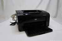 Front-right angled view of a black HP LaserJet Pro P1102w printer with input/output trays open, showing clean exterior and cables attached.