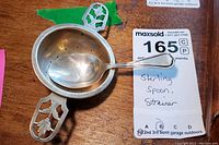 Photo shows the sterling spoon resting inside the sterling strainer on a wooden surface with the lot tag visible.
