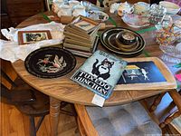 Table displaying four Fifi and Pepe decorative trays, stone welcome sign, black enamelware wall pocket dish, and several framed prints stacked together.