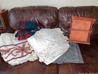 Photo of multiple vintage bedspreads and blankets including Scottish mohair blanket, U.S. military wool blanket, and a wooden display shelf on a dark brown leather couch.